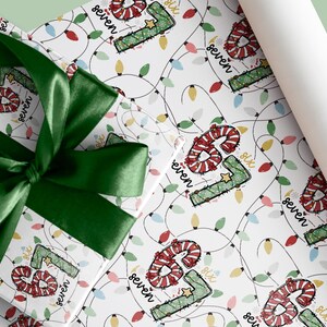 Pode incluir: Um presente embrulhado com um laço de cetim verde está sobre papel de embrulho a condizer. O papel apresenta um padrão festivo de luzes de Natal e os números "6" e "seven" num design de bengala doce.