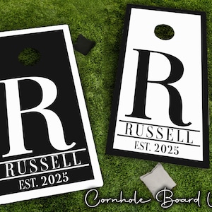 Könnte beinhalten: Zwei weiße Cornhole-Bretter mit schwarzem Rand und einem schwarzen Buchstaben "R" mit dem Namen "Russell" und "Est. 2025" aufgedruckt.