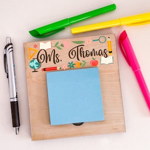 Puede incluir: Un portanotas de madera con el nombre "Ms. Thomas" e ilustraciones escolares. Un bloc de notas adhesivas azul está adjunto. Un bolígrafo plateado y tres rotuladores de colores verde, amarillo y rosa están colocados alrededor del portanotas.