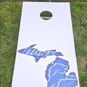 A white cornhole board with a blue lion silhouette and the word "LIONS" printed on it, placed on a grassy surface.