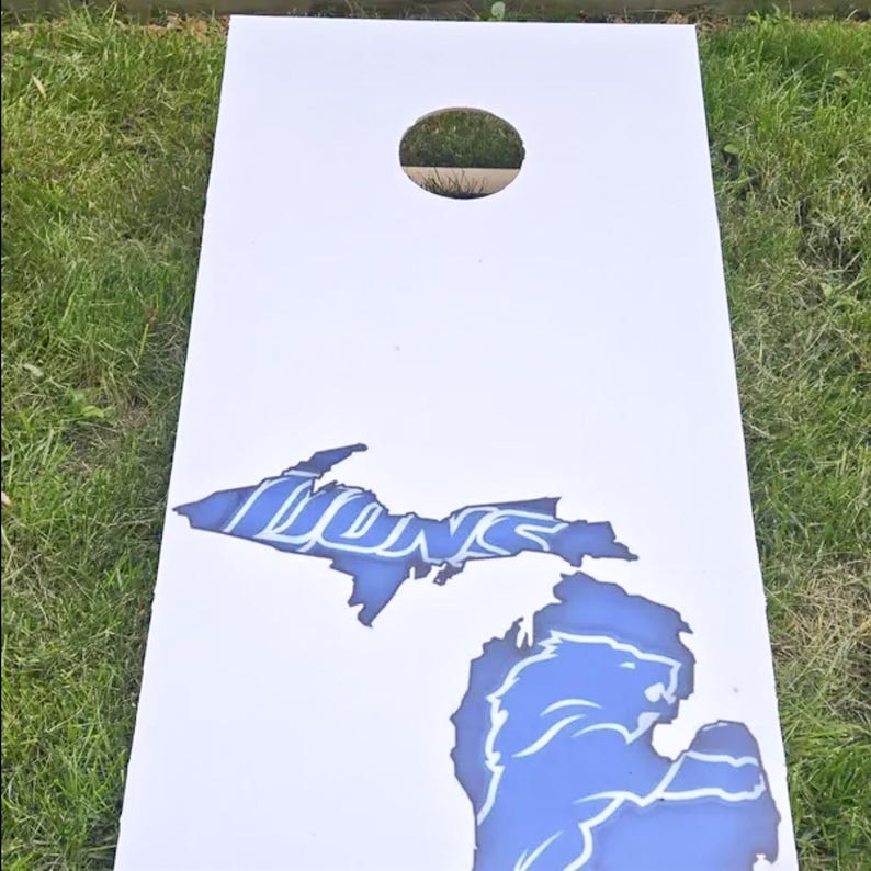 A white cornhole board with a blue lion silhouette and the word "LIONS" printed on it, placed on a grassy surface.