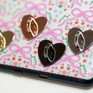 May include: A close-up of a phone case with a floral and bow pattern. Four heart-shaped phone grips in gold, rose gold, and silver are attached to the case. The case is black.