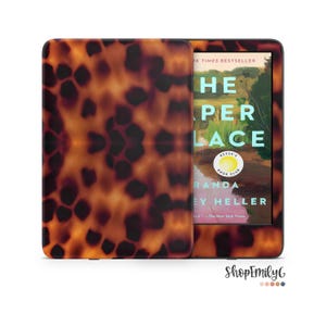Op de afbeelding: Een e-reader met een schildpadpatroon, open om de omslag van een boek met de titel "The Keeper Place" van Randa Heller te tonen. De omslag heeft een landschapsscène met groene en bruine tinten.