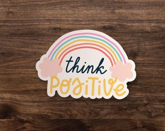 May include: A die-cut sticker featuring a pastel rainbow arching over two light pink clouds. The word think is written in dark blue script, and the word POSITIVE is written in large, bold yellow letters below the rainbow and clouds.