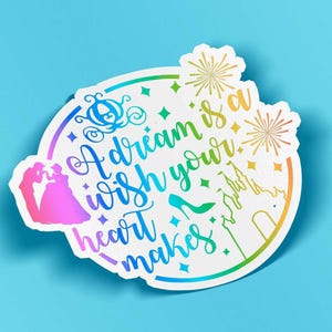 May include: A circular sticker with a white background featuring the text A dream is a wish your heart makes in a rainbow gradient. The design includes a silhouette of a couple, a carriage, a glass slipper, a castle, and fireworks icons.