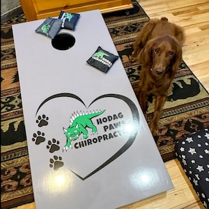 A brown dog is sitting on a rug next to a gray cornhole board with a green dinosaur and paw prints, and a small black bag with a white logo.