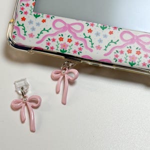 Op de afbeelding: Een doorzichtige telefoonhoes met een bloemen- en strikpatroon. Twee roze strikbedels met parelaccenten liggen naast de hoes. De hoes heeft een witte achtergrond met roze strikken, rode en blauwe bloemen en groene ranken.
