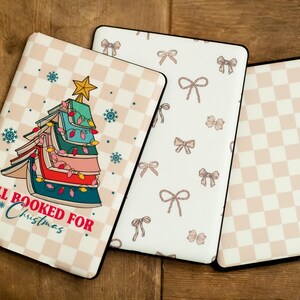 Op de afbeelding: Drie verschillende patroonhoezen voor een e-reader. De eerste hoes heeft een kerstboom gemaakt van boeken met de tekst "All Booked For Christmas". De tweede hoes heeft een witte achtergrond met een herhalend patroon van roze strikken. De derde hoes heeft een witte achtergrond met een herhalend patroon van roze en witte vierkanten.