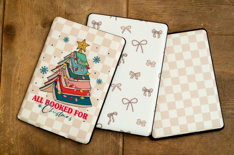Op de afbeelding: Drie verschillende patroonhoezen voor een e-reader. De eerste hoes heeft een kerstboom gemaakt van boeken met de tekst "All Booked For Christmas". De tweede hoes heeft een witte achtergrond met een herhalend patroon van roze strikken. De derde hoes heeft een witte achtergrond met een herhalend patroon van roze en witte vierkanten.