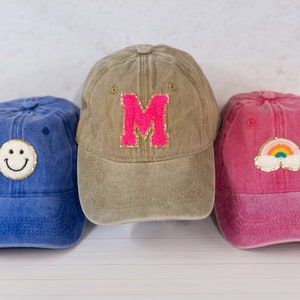Op de afbeelding: Drie denim baseballpetten met verschillende geborduurde patches. De eerste pet is blauw met een gele smiley, de tweede is kaki met een roze letter M en de derde is roze met een regenboog en een wolk.
