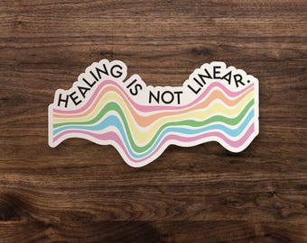 May include: A die-cut sticker featuring the black text "HEALING IS NOT LINEAR." positioned above a series of wavy, horizontal lines in pastel shades of pink, yellow, green, and blue, creating a fluid, multi-colored graphic design.
