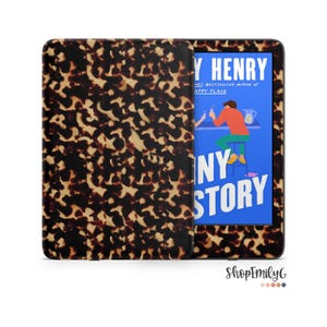 May include: An e-reader device featuring a protective case with a brown and black tortoiseshell pattern. The screen displays the book cover for Funny Story by Emily Henry, showing a blue background with a person sitting on a stool at a bar.