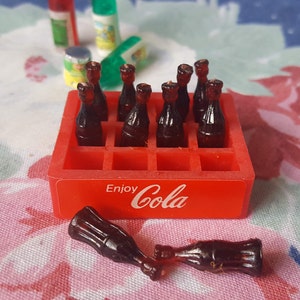 May include: A red plastic crate with the words "Enjoy Cola" printed on the front. The crate is filled with miniature glass bottles of cola. Two bottles are lying outside the crate.