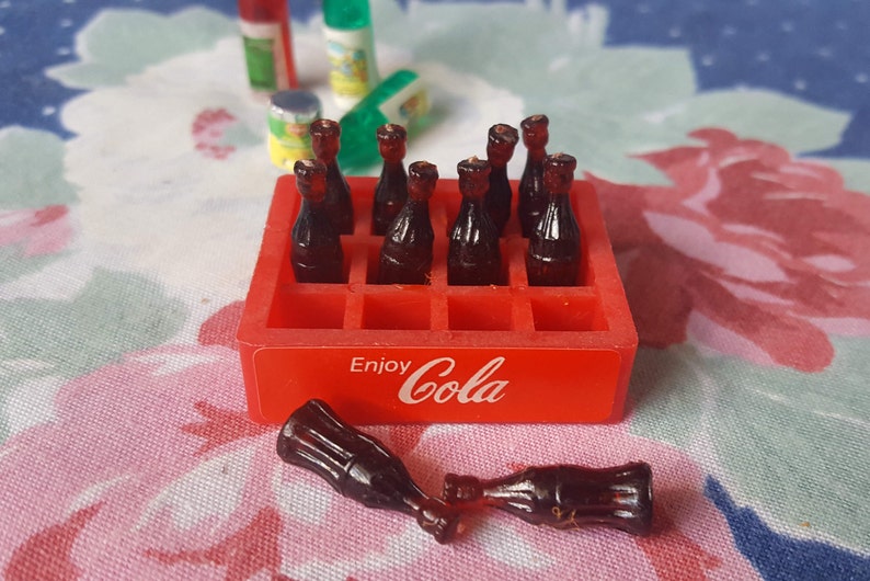 May include: A red plastic crate with the words "Enjoy Cola" printed on the front. The crate is filled with miniature glass bottles of cola. Two bottles are lying outside the crate.