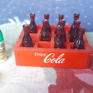 May include: A red plastic crate with the words "Enjoy Cola" printed on the front. The crate is filled with miniature brown glass bottles.