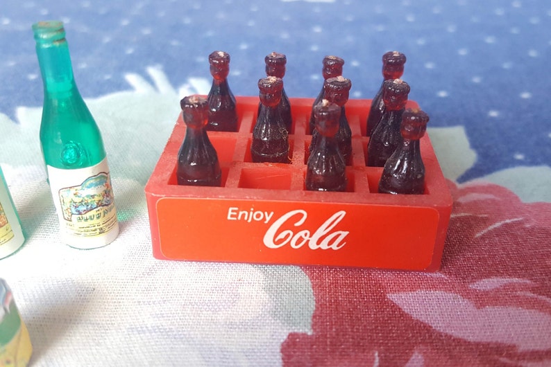 May include: A red plastic crate with the words "Enjoy Cola" printed on the front. The crate is filled with miniature brown glass bottles.