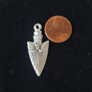 May include: A silver-colored arrowhead pendant with a rope-like detail around the base. The pendant is about the size of a US penny.