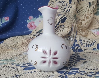 May include: White glass pitcher with a clear handle and a flared rim. The pitcher features a decorative floral design in the center and gold accents. The rim and the center of the floral design are a deep pink color. The pitcher is a decorative item.