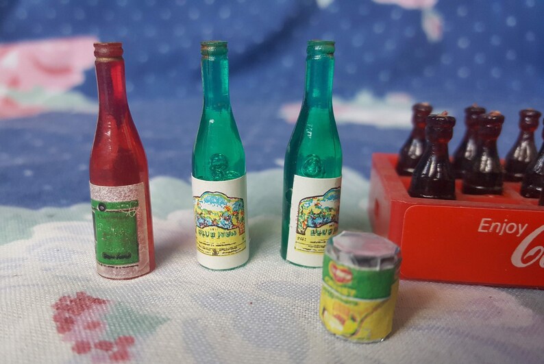 May include: A miniature set of toy bottles, including two green glass wine bottles with labels, a red glass wine bottle with a label, and a six-pack of brown glass soda bottles in a red and white cardboard box with the word "Enjoy" printed on it. There is also a small green and yellow can with a label.