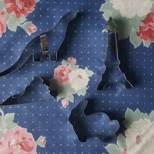 May include: Four metal cookie cutters on a blue and white floral fabric background. The cutters are shaped like a dinosaur, a rabbit, a mountain, and the Eiffel Tower.