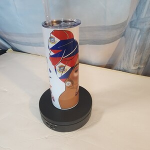 May include: A white stainless steel tumbler with a design of a woman wearing a headscarf with the Puerto Rican flag colors and a gold earring. The tumbler is on a black rotating display stand.