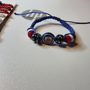 May include: A blue braided bracelet featuring red, white, and black beads. The center bead displays the Haitian flag. The bracelet is adjustable with a sliding knot closure. Other bracelets are visible in the background.