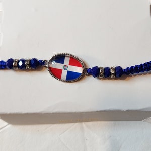 May include: A blue and white string bracelet with a silver oval charm featuring the Dominican Republic flag. The charm has a clear glass covering over the flag.