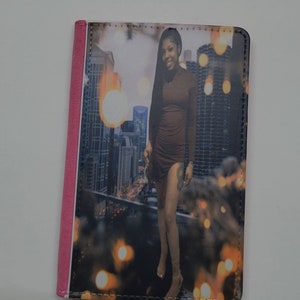 May include: A black leather passport holder with a pink edge. The front of the holder features a photo of a person standing in front of a city skyline.