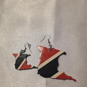 May include: A pair of dangling earrings shaped like the island of Trinidad, featuring the national flag colors of red, black, and white with a diagonal stripe. The earrings are attached to silver-toned metal hooks with clear plastic backings.