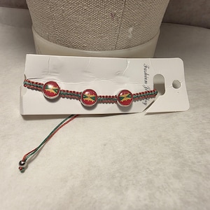 Może przedstawiać: Czerwona, zielona i żółta bransoletka z trzema okrągłymi zawieszkami z wzorem. Bransoletka wykonana jest z plecionych czerwonych i zielonych nici. Prezentowana jest na białej karcie z napisem "Fashion Jewelry".