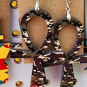 May include: A pair of earrings featuring a camouflage pattern Ankh design. The earrings are made of wood and have silver hooks.