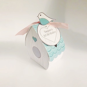 Puede incluir: Una caja de regalo de pájaro blanco y azul con una cinta rosa y una etiqueta en forma de corazón que dice "Tegan's 1st Birthday".