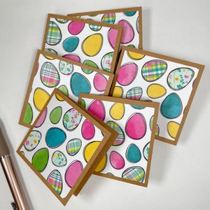 Könnte beinhalten: Ein Stapel quadratischer Grußkarten mit braunem Rand. Die Karten zeigen einen weißen Hintergrund mit bunten Osterei-Designs in Pink, Gelb, Türkis und Grün mit verschiedenen Mustern. Ein Stift ist in der Ecke sichtbar.
