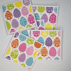 May include: Four square Easter-themed coasters with colorful egg designs. Each coaster features a white border and a pattern of decorated eggs in various colors, including pink, purple, yellow, and green. One coaster has the text "happy easter".