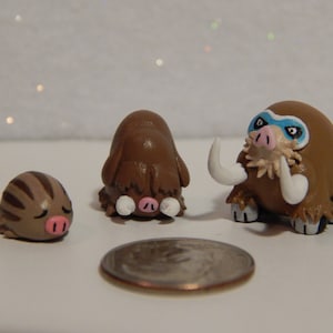 May include: Three handmade clay figurines of Pokemon: a brown and white Swinub, a brown and white Piloswine, and a brown and white Mamoswine. The Mamoswine has blue eyes and a white mask.