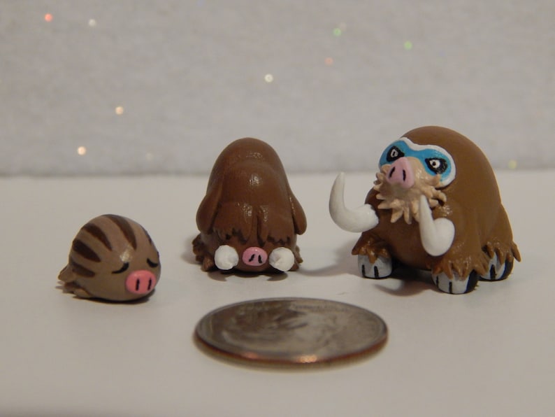 May include: Three handmade clay figurines of Pokemon: a brown and white Swinub, a brown and white Piloswine, and a brown and white Mamoswine. The Mamoswine has blue eyes and a white mask.