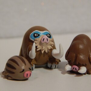 May include: Three handmade clay figurines of brown and white Pokemon. The largest figurine is a brown and white Mamoswine with a blue mask. The middle figurine is a brown and white Pignite. The smallest figurine is a brown and white Swinub.
