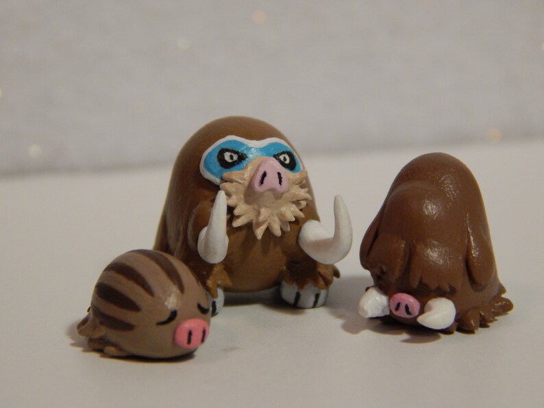 May include: Three handmade clay figurines of brown and white Pokemon. The largest figurine is a brown and white Mamoswine with a blue mask. The middle figurine is a brown and white Pignite. The smallest figurine is a brown and white Swinub.