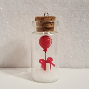 Peut inclure: Une petite bouteille en verre avec un bouchon en li&egrave;ge contient un ballon rouge attach&eacute; &agrave; une bo&icirc;te cadeau blanche avec un ruban rouge. La bouteille est remplie de paillettes blanches.