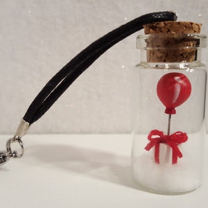 Peut inclure: Une petite bouteille en verre avec un bouchon en li&egrave;ge contient un ballon rouge attach&eacute; &agrave; une petite bo&icirc;te cadeau blanche avec un n&oelig;ud rouge. La bouteille est attach&eacute;e &agrave; un cordon noir avec un fermoir argent&eacute;.