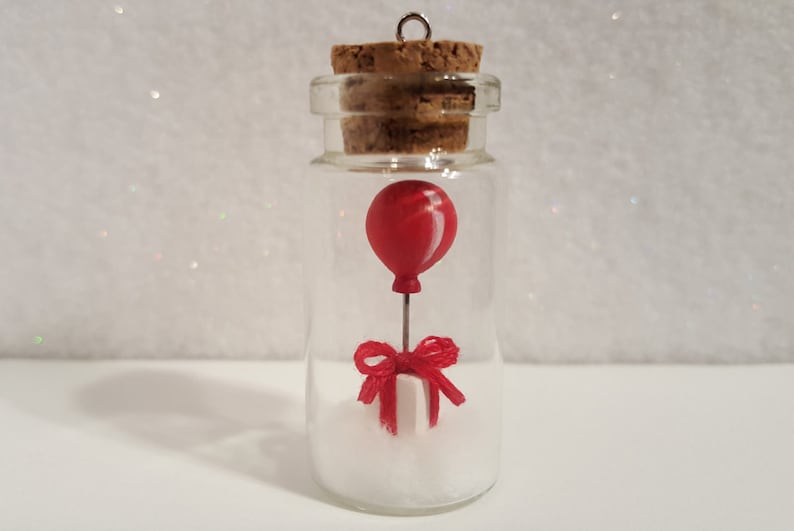 Peut inclure: Une petite bouteille en verre avec un bouchon en li&egrave;ge contient un ballon rouge attach&eacute; &agrave; une bo&icirc;te cadeau blanche avec un n&oelig;ud rouge. La bouteille est remplie de paillettes blanches.