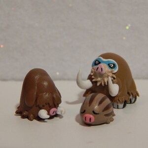 May include: Three brown and white clay figurines of Pokemon characters. The largest figurine is a brown and white Mamoswine with blue eyes and a white mask. The middle figurine is a brown and white Swinub with a pink nose. The smallest figurine is a brown and white Piloswine with a pink nose.