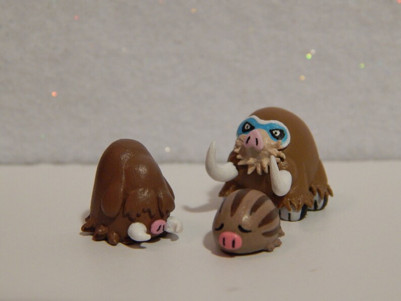 May include: Three brown and white clay figurines of Pokemon characters. The largest figurine is a brown and white Mamoswine with blue eyes and a white mask. The middle figurine is a brown and white Swinub with a pink nose. The smallest figurine is a brown and white Piloswine with a pink nose.