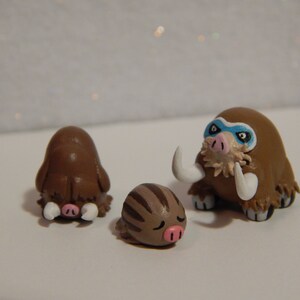 May include: Three handmade clay figurines of brown and white woolly mammoths. The largest mammoth has a blue mask painted on its face.