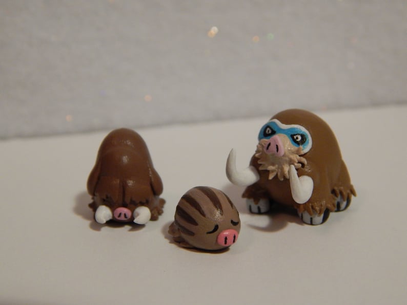 May include: Three handmade clay figurines of brown and white woolly mammoths. The largest mammoth has a blue mask painted on its face.