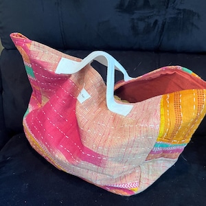 May include: A tote bag constructed from patchwork fabric in shades of pink, orange, and yellow, featuring white stitched detailing throughout the surface. The bag includes two white fabric handles and a small white rectangular label on the front.