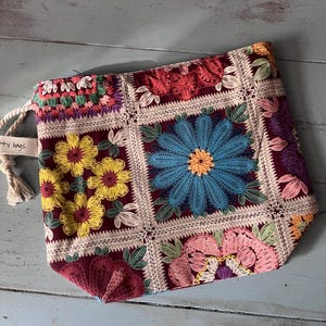 Può includere: Una pochette con coulisse caratterizzata da un design patchwork di quadrati floreali ricamati su uno sfondo bordeaux intenso. Il ricamo include vivaci fiori gialli, blu e rosa con foglie verdi. Una piccola etichetta bianca riporta la scritta "ty bags".