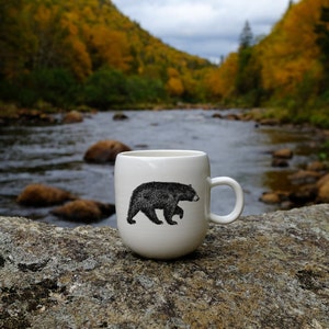 Può includere: Una tazza in ceramica bianca con un design a silhouette di orso nero si trova su una roccia grigia vicino a un fiume. Lo sfondo è un'immagine sfocata di una foresta con foglie autunnali.