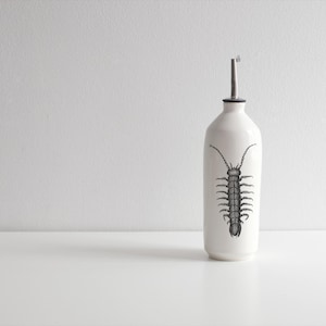 Peut inclure: Bouteille d'huile en céramique blanche avec une illustration de mille-pattes noire. La bouteille a un bec verseur argenté et est posée sur une surface blanche. Design minimaliste et moderne.