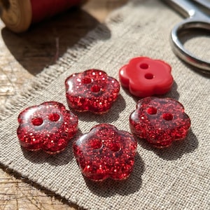 May include: Four flower-shaped buttons with a red glitter finish and one solid red flower-shaped button, each featuring two central holes. These five buttons are arranged on a piece of light beige fabric near a spool of red thread and scissors.